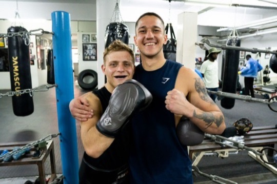 Two boxers smile with their arms around each other beside heavy bags.