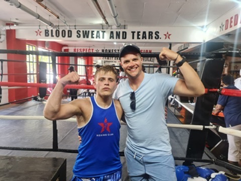 Two boxers smile together in front of a heavy bag.